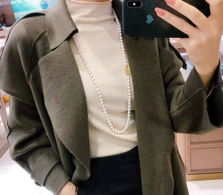 Person wearing a green coat over a white top with a pearl necklace, taking a mirror selfie.