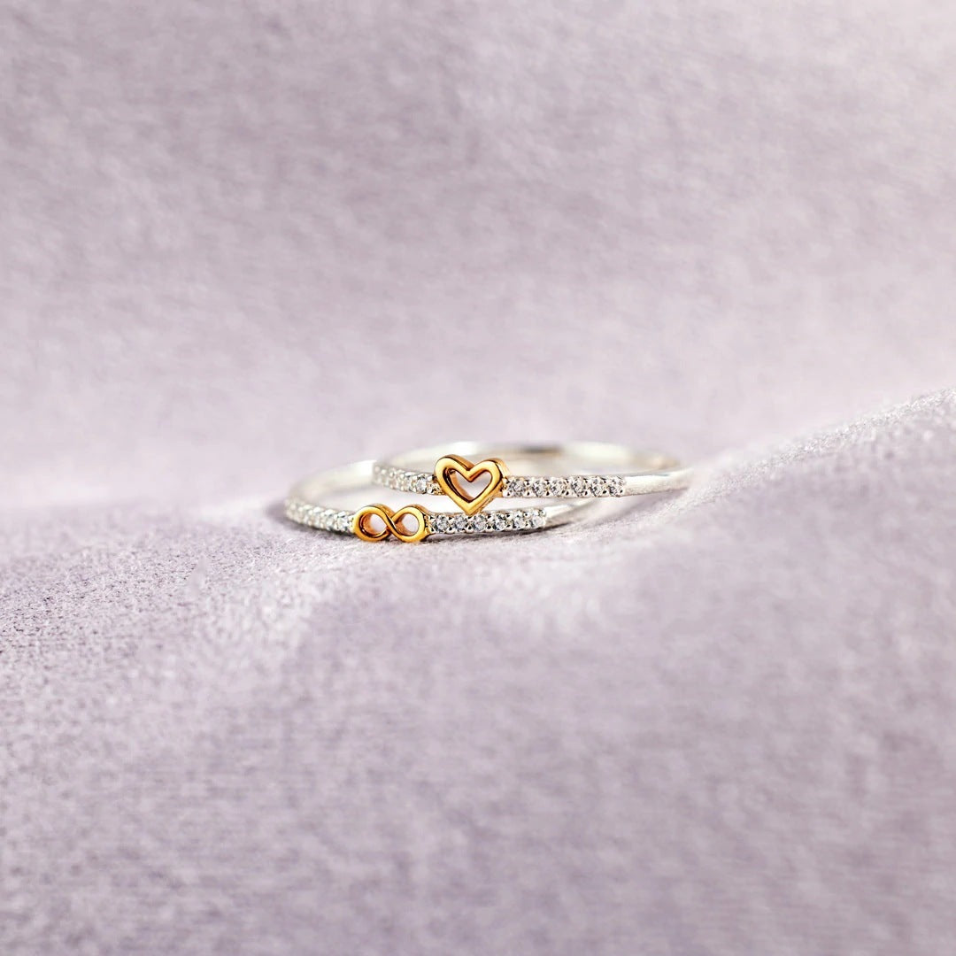 Two rings with heart and infinity symbol designs on a light purple fabric background