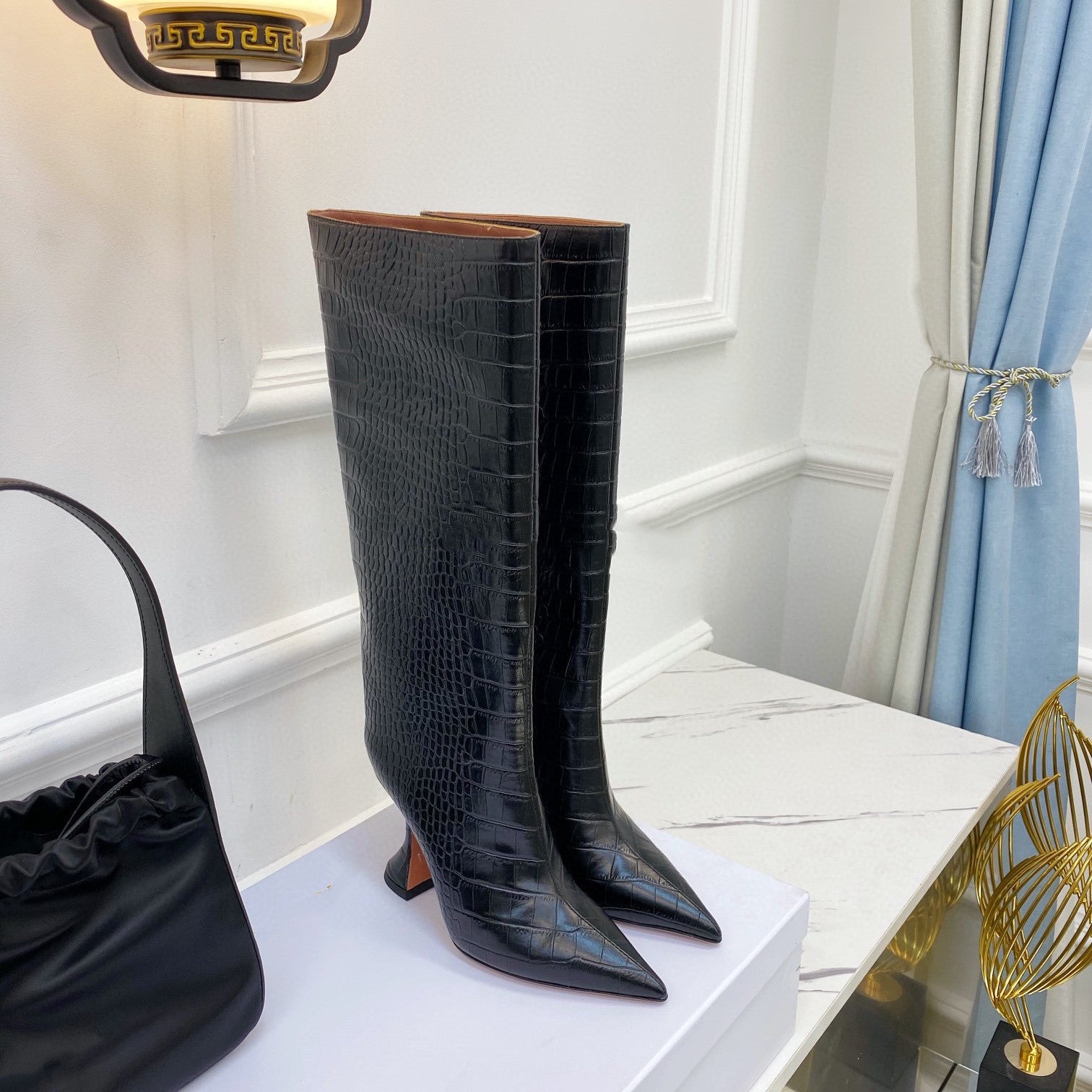 Black crocodile-patterned knee-high boots on a white surface with a black handbag and decorative item in the background.