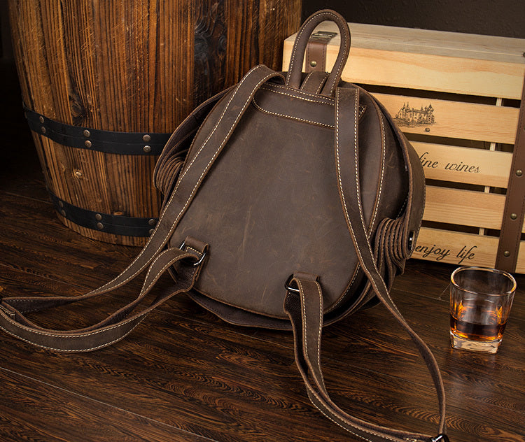 Brown leather backpack on a wooden surface with a glass of whiskey and wooden barrel in the background.