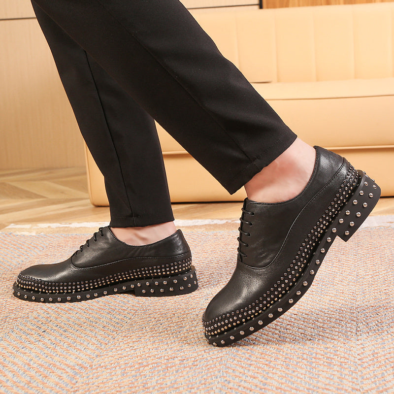 Black leather shoes with studs worn by a person on a textured floor.