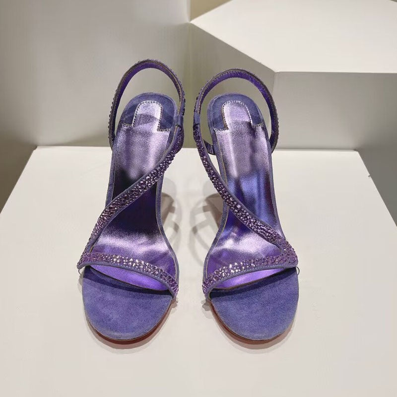 Pair of purple high-heeled sandals with embellishments on a white surface.