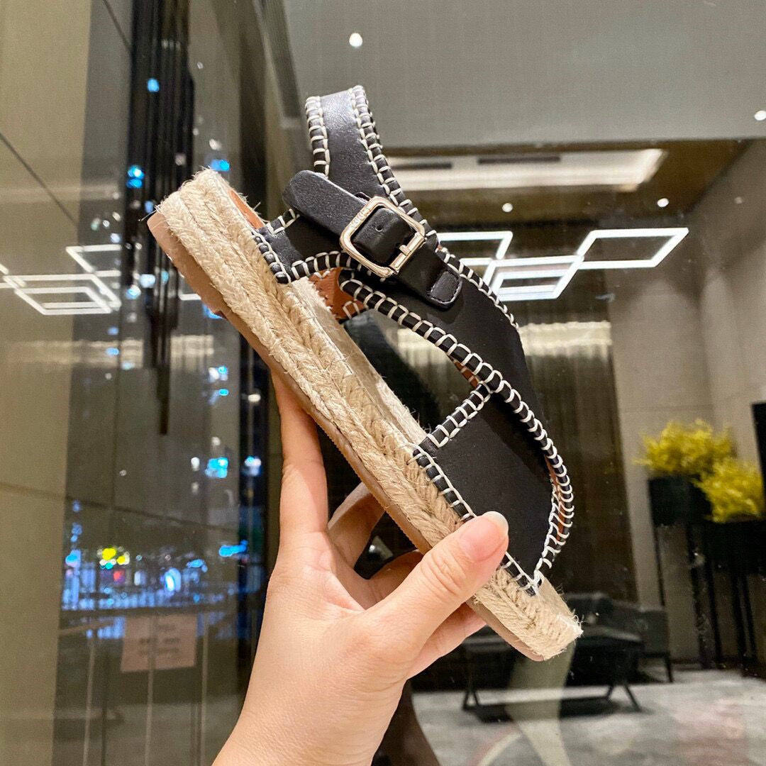 Hand holding a sandal with a black strap and beige sole in front of a glass door.