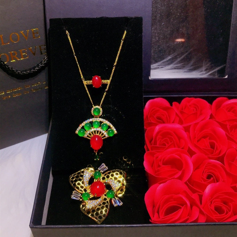 Gold necklace with red and green gemstones on a black card, next to red roses.