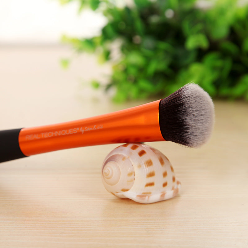 Makeup brush with orange handle and black bristles on a shell against a blurred green plant background