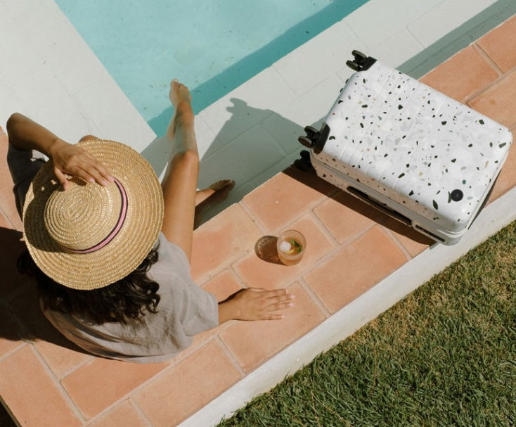 Person sitting by a pool with a speckled suitcase and straw hat