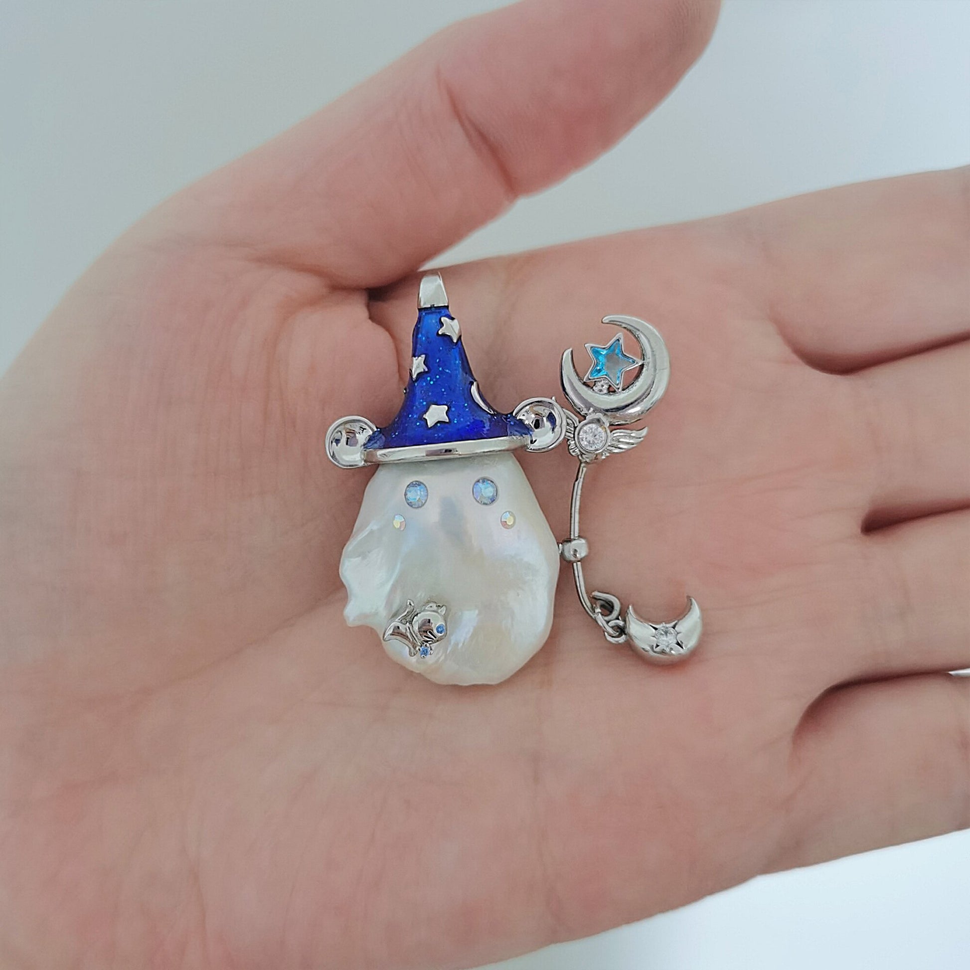 Decorative brooch with a blue hat, star, moon, and pearl-like center held in a hand.