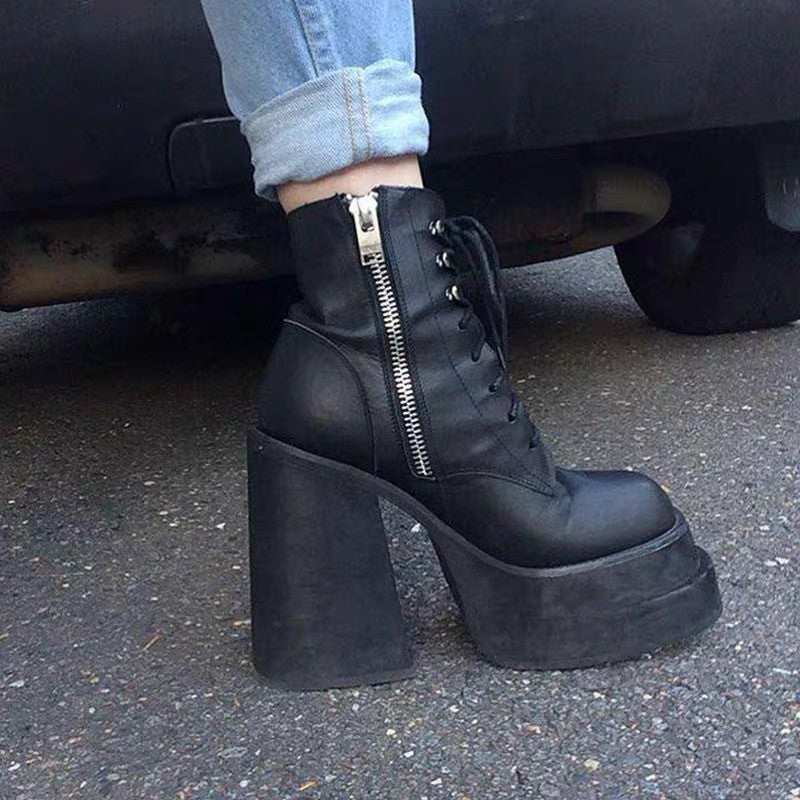 Black platform boots with a high heel worn by a person, with a car in the background.