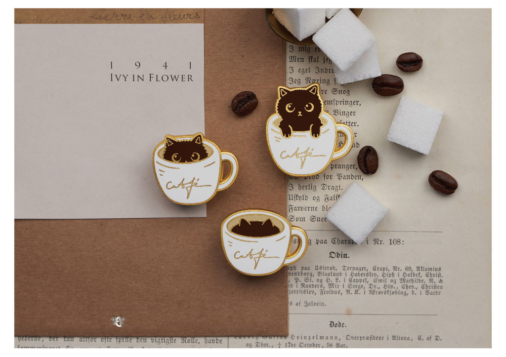 Three decorative pins featuring cats in coffee cups on a textured surface with coffee beans and marshmallows.