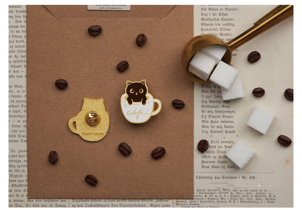 Two enamel pins on a surface with coffee beans and a spoon.