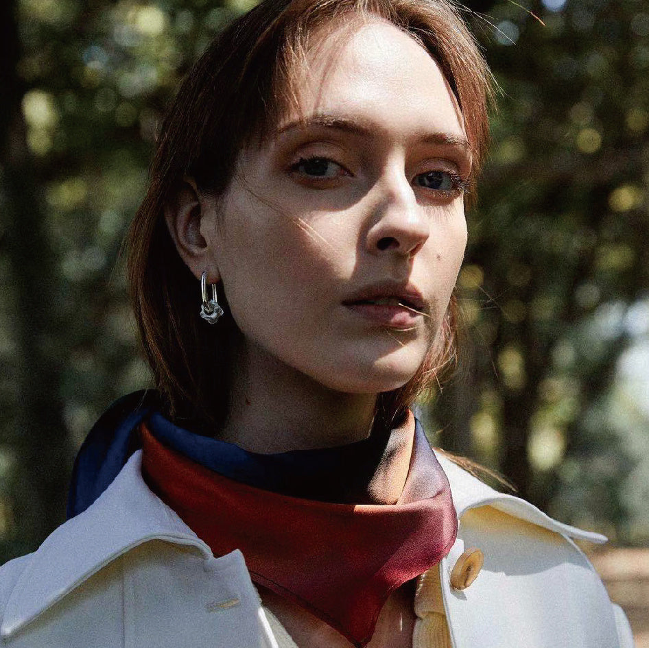 Woman wearing a white coat with a red scarf, standing outdoors with trees in the background