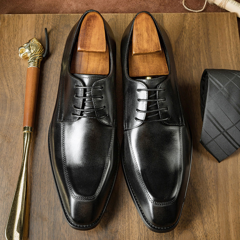Pair of black dress shoes on a wooden surface with a gold shoe horn and black bag.