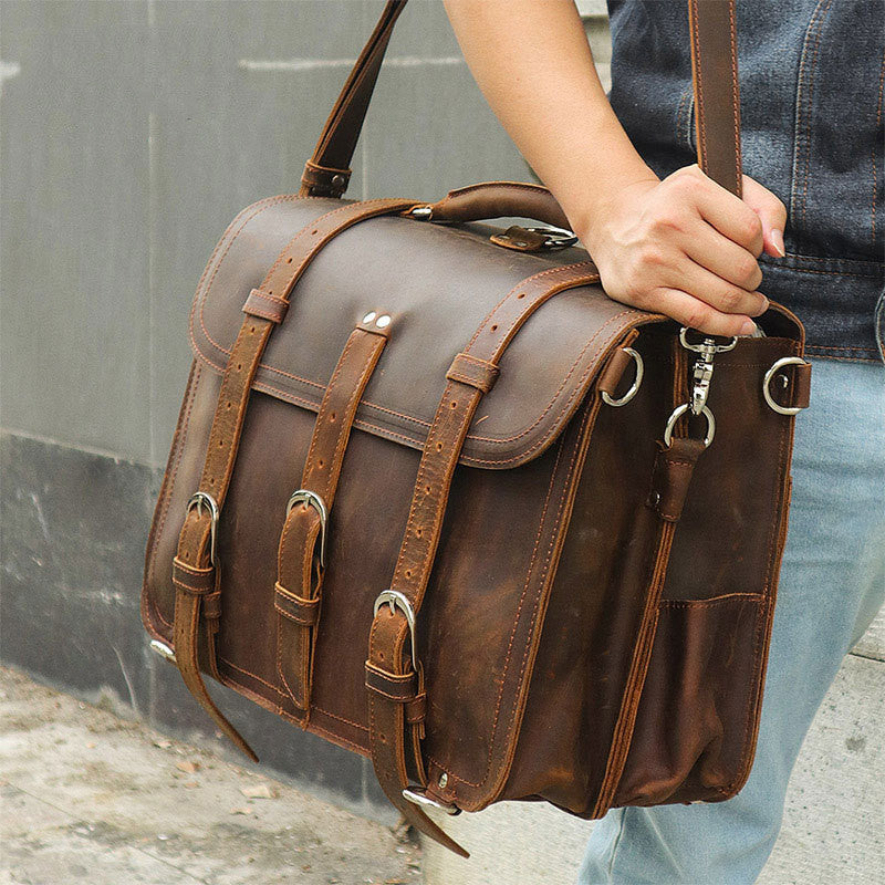 Brown leather satchel bag held by a person against a gray wall.