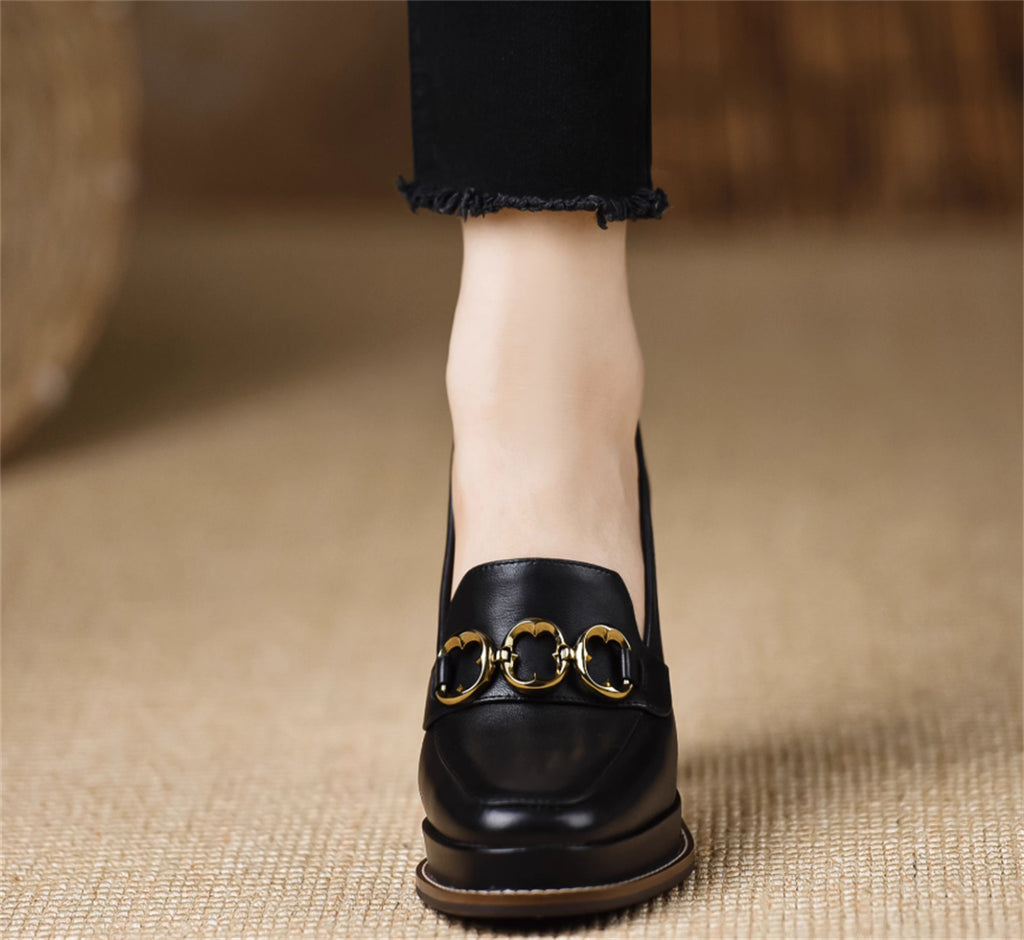 Black loafer with gold buckle worn by a person on a textured beige surface