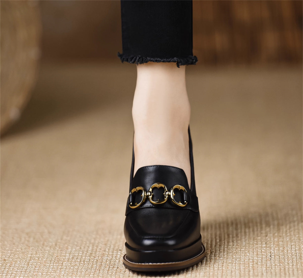Black loafer with gold buckle worn by a person on a textured beige surface