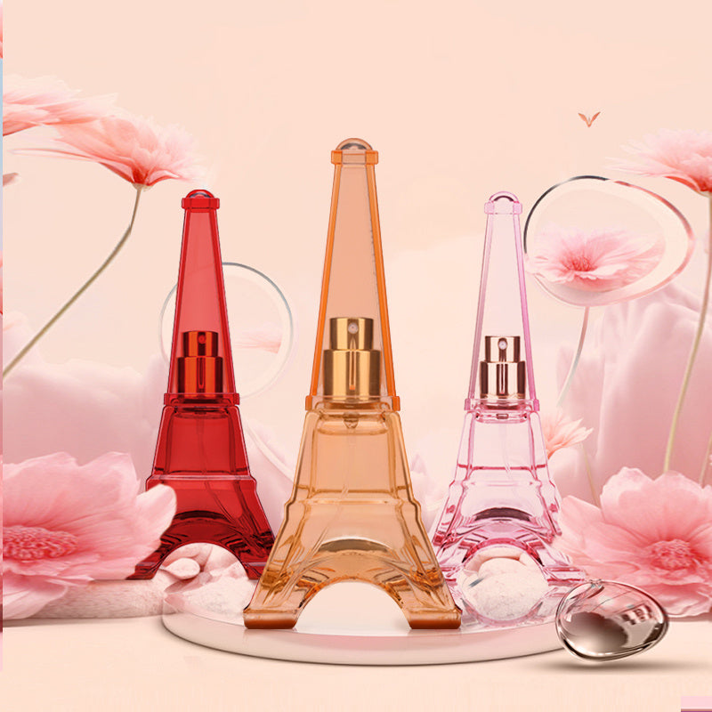Three Eiffel Tower-shaped perfume bottles in red, orange, and pink on a decorative stand with flowers in the background.