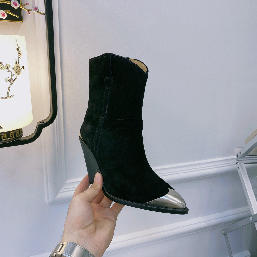 Black suede ankle boot with metallic heel held by a hand against a white wall.