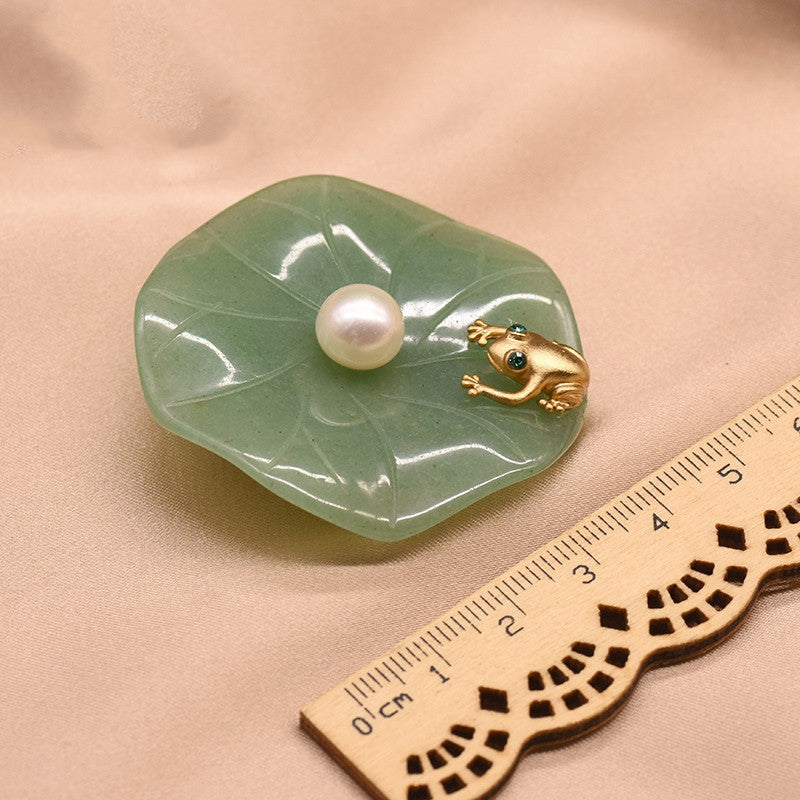 Green jade dish with a pearl and gold frog on a beige background, accompanied by a wooden ruler for scale.