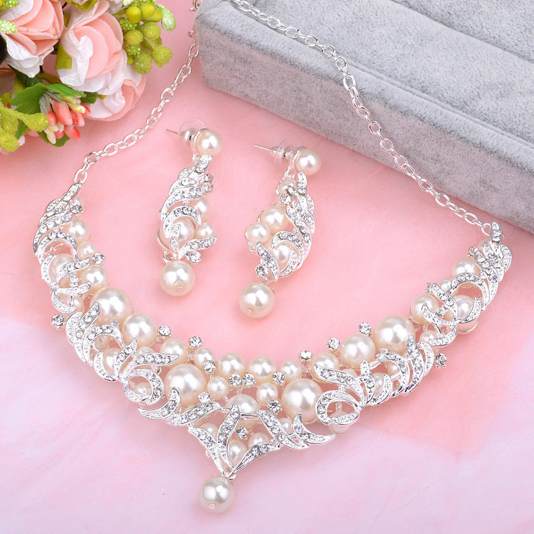 Pearl necklace and earrings set on a pink surface with flowers and jewelry box in the background.