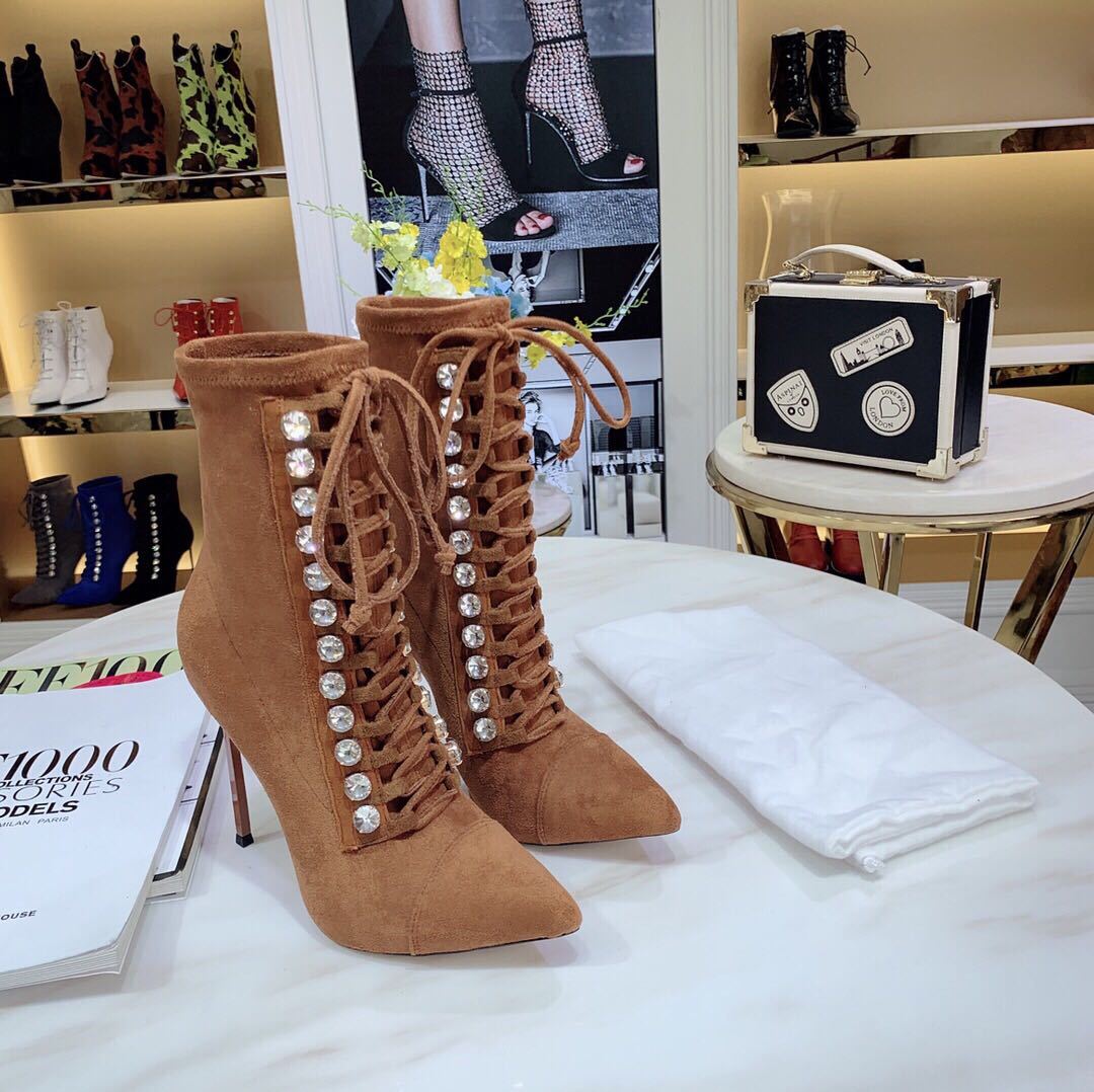 Brown suede high-heeled boots with embellishments on a white surface, with a magazine and small black suitcase in the background.