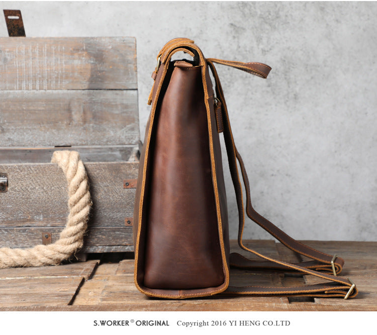 Brown leather bag with strap on a wooden surface against a gray wall.