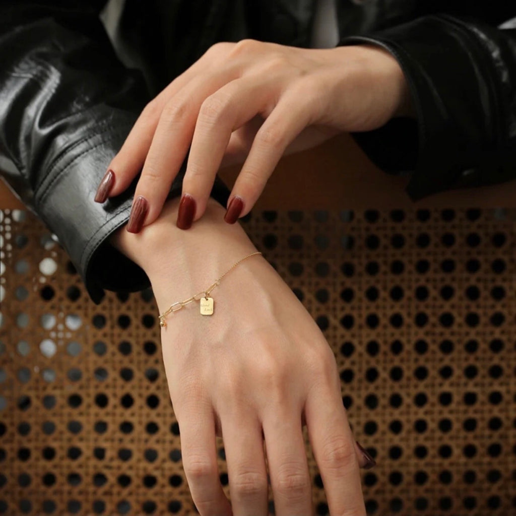 Close-up of a hand wearing a gold bracelet with a small tag, against a textured brown background.
