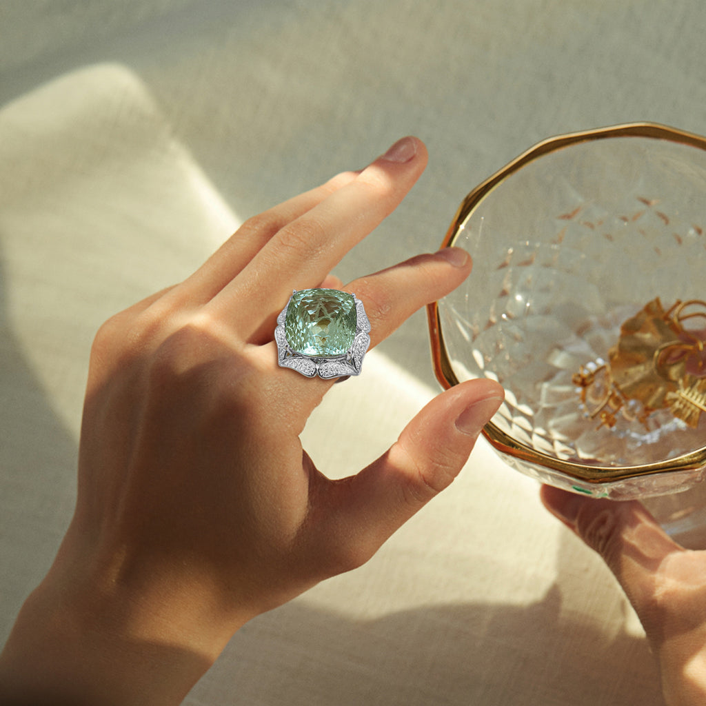 Hand wearing a large green gemstone ring holding a glass dish with gold leaf.
