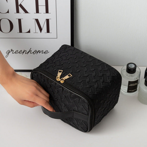 Black textured toiletry bag with gold zipper on a white surface, with a hand reaching towards it.