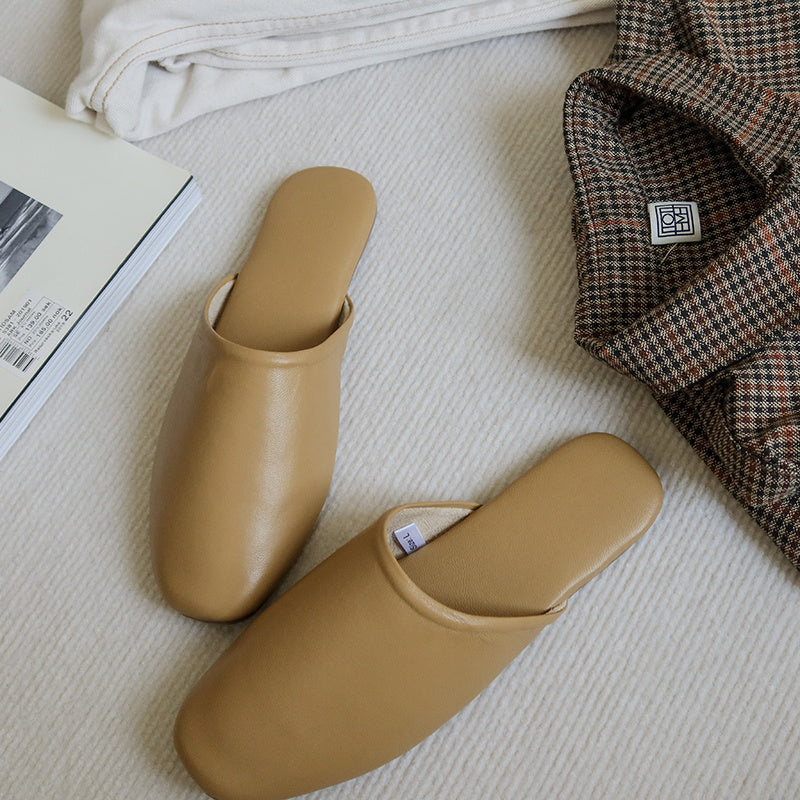 Pair of beige slippers on a textured surface with a plaid garment and newspaper in the background.