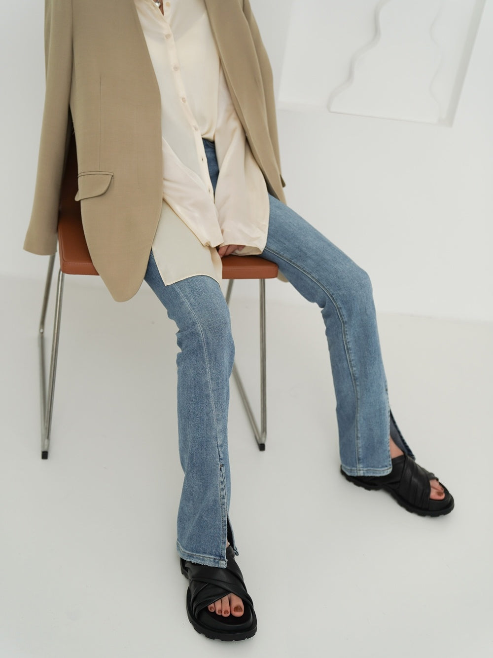Person wearing a beige coat, white shirt, blue jeans, and black sandals sitting on a chair.