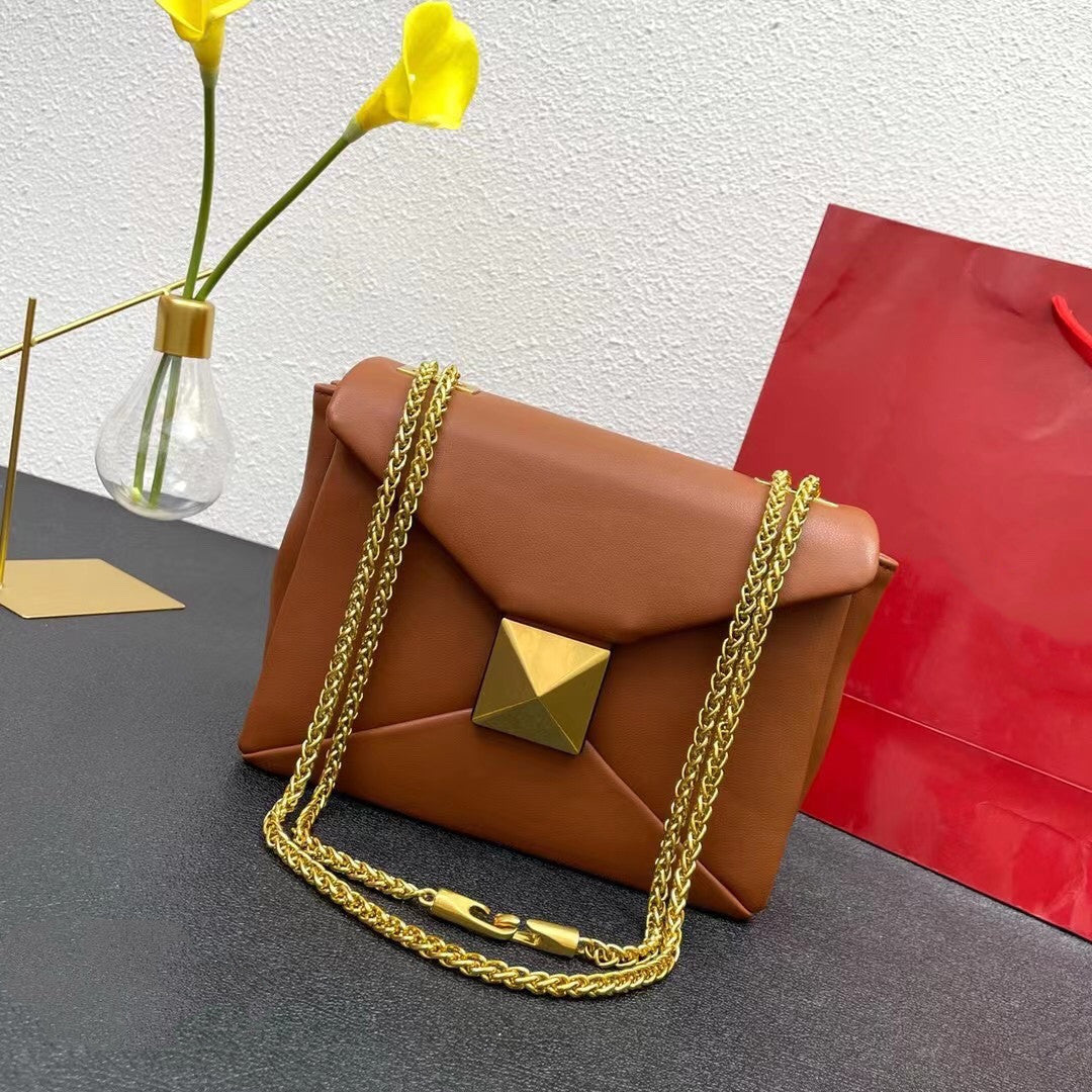 Brown handbag with gold chain and clasp on a gray surface with a red bag and yellow flowers in the background.