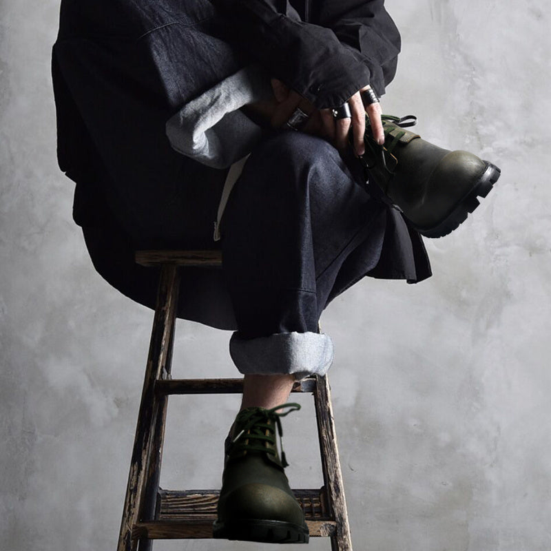 Person sitting on a wooden stool wearing dark clothing and green boots against a gray background