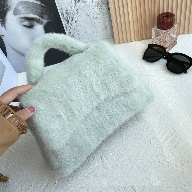 Light green fluffy clutch held by a hand on a white surface with sunglasses and a bottle in the background.