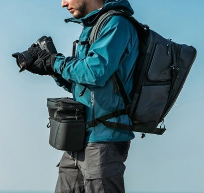 Person in teal jacket with backpack and camera equipment against a clear blue sky