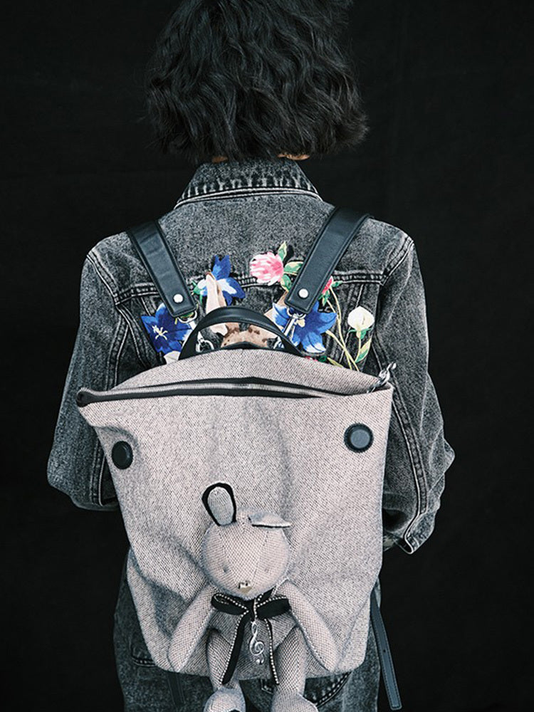 Person wearing a denim jacket with floral embroidery, carrying a backpack with a plush toy on a black background