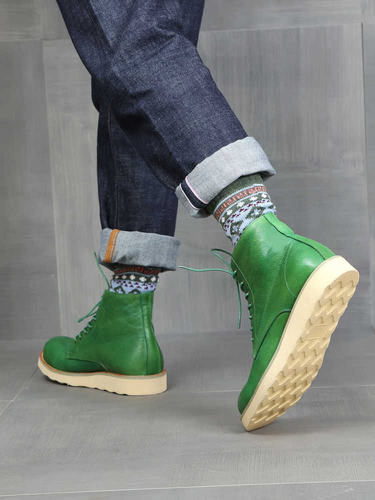 Green leather boots with beige soles worn with patterned socks and rolled-up jeans on a gray floor.