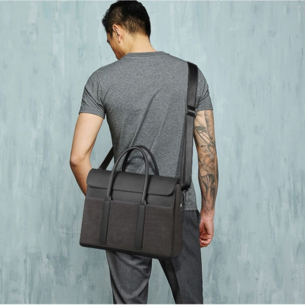 Man carrying a dark gray leather briefcase against a light blue wall.