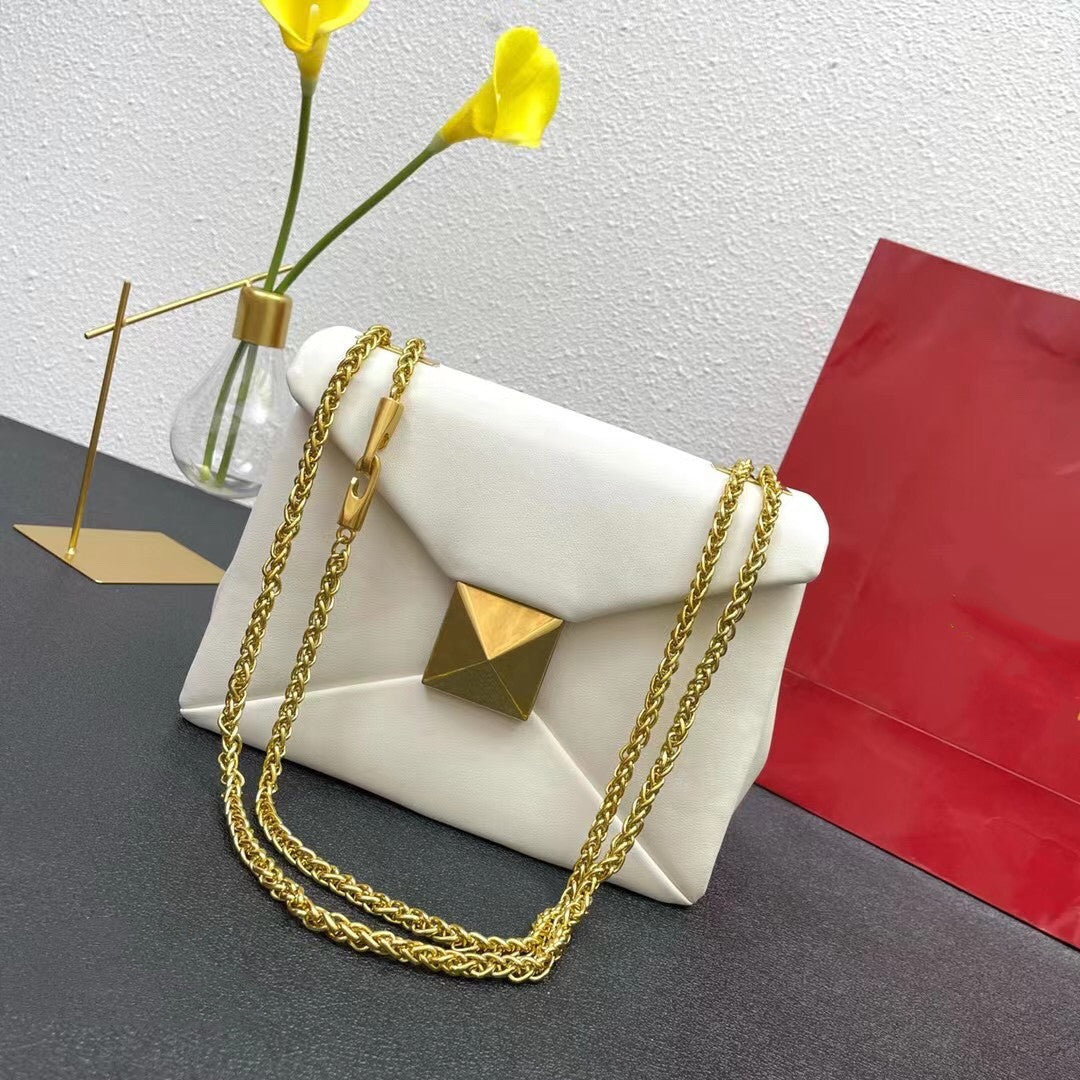 White handbag with gold chain and geometric design on a gray surface with a red bag and yellow flowers in the background.