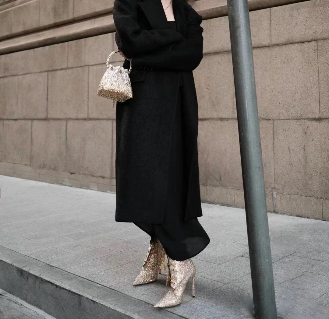 Person wearing a black outfit with high-waisted pants and silver shoes, standing against a concrete wall.