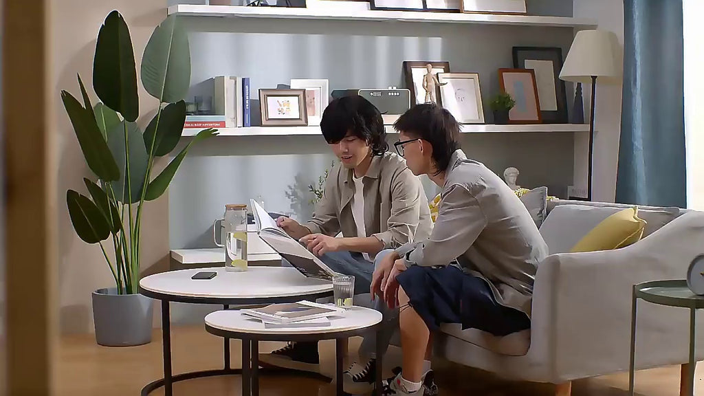 Two people sitting on a couch in a modern living room with a plant and books on shelves.
