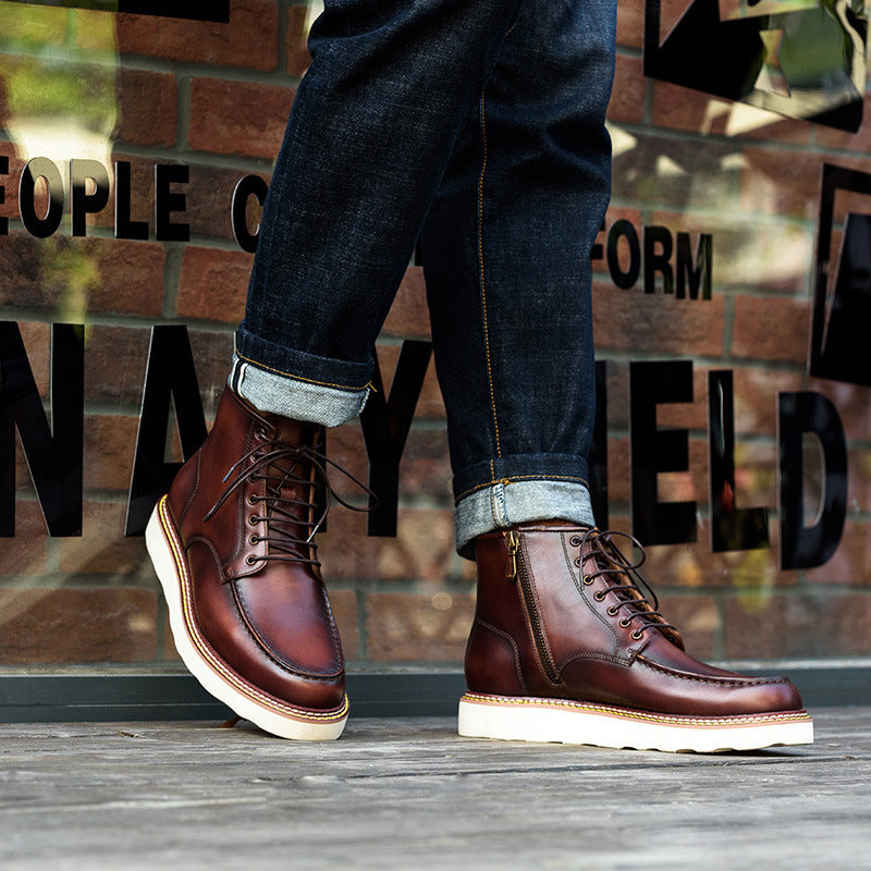 Brown leather boots with white soles worn with blue jeans against a brick wall.