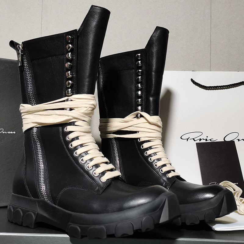 Black leather boots with white laces on a gray surface with a branded paper bag in the background.