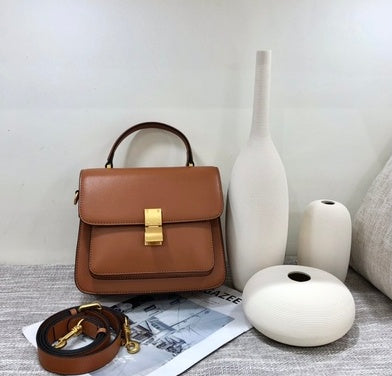 Brown handbag with gold clasp on a surface with white vases and newspaper.