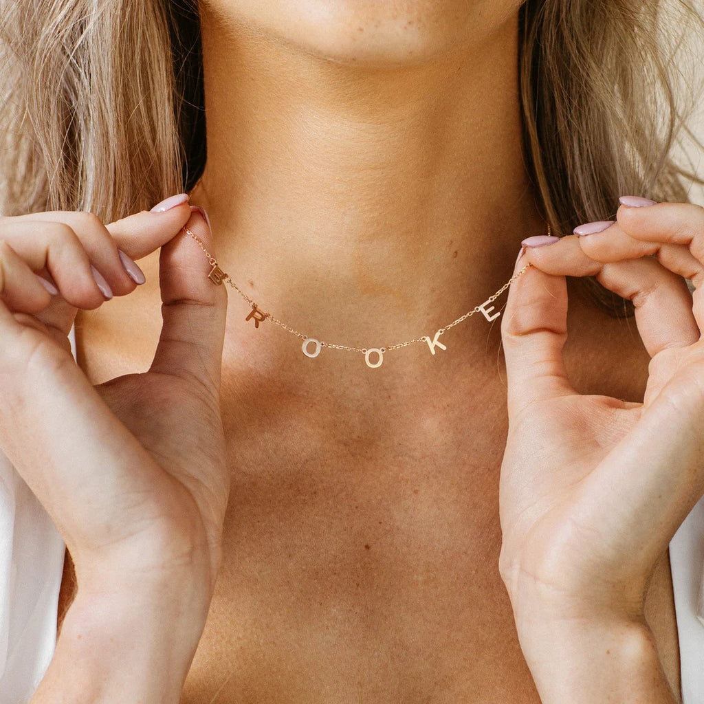 Person wearing a gold necklace with letter charms, held by hands.