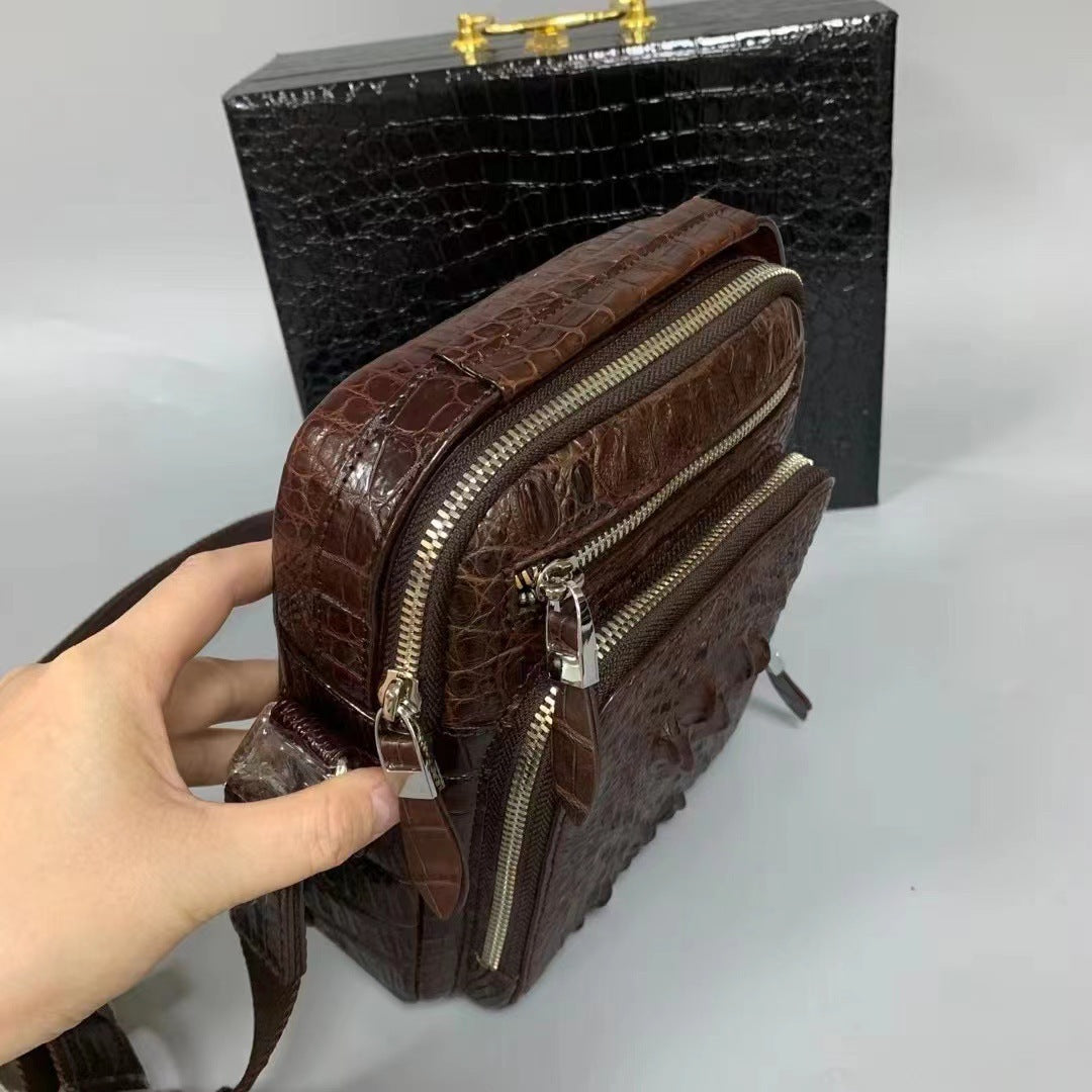 Brown crocodile-patterned handbag with gold zippers held by a hand, against a black textured box on a white background.