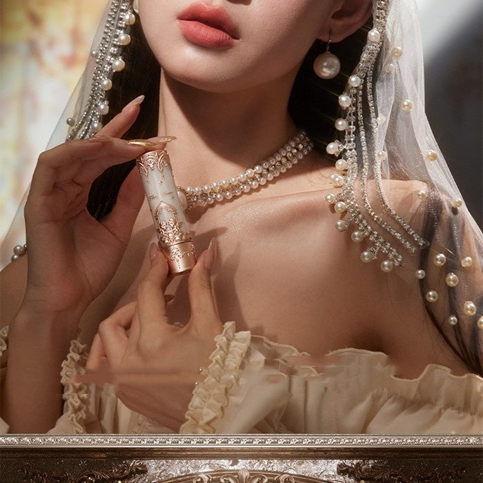 Close-up of a woman holding a decorative lipstick with intricate details, wearing a pearl necklace and earrings.