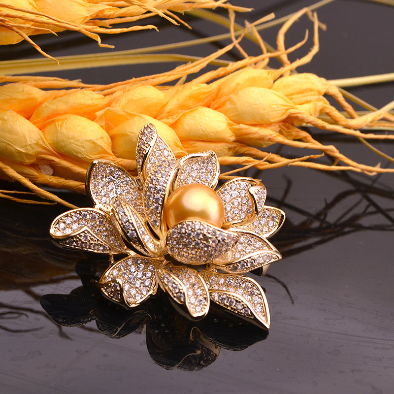 Gold flower-shaped brooch with a pearl center on a reflective surface with yellow flowers in the background.