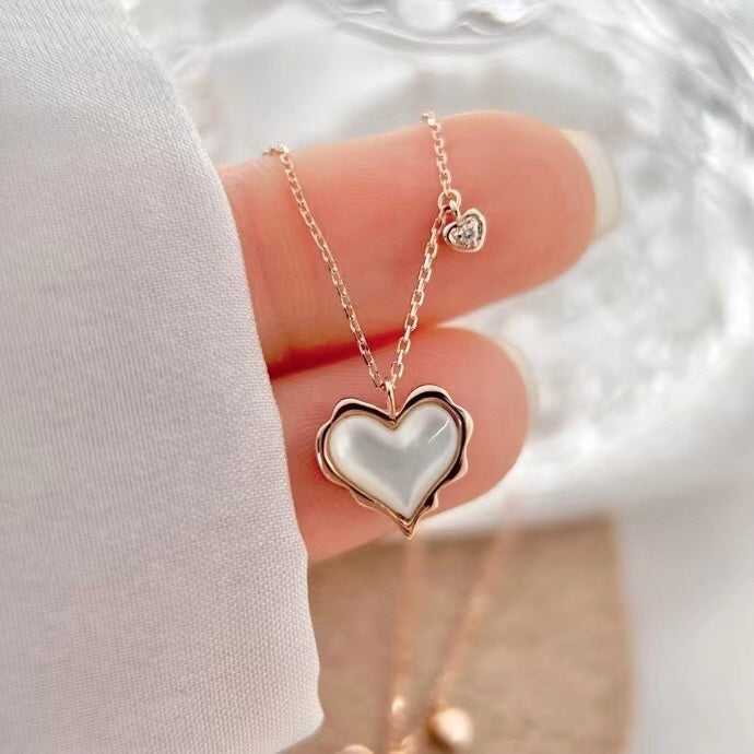 Heart-shaped necklace with a white heart pendant held by a hand against a white background