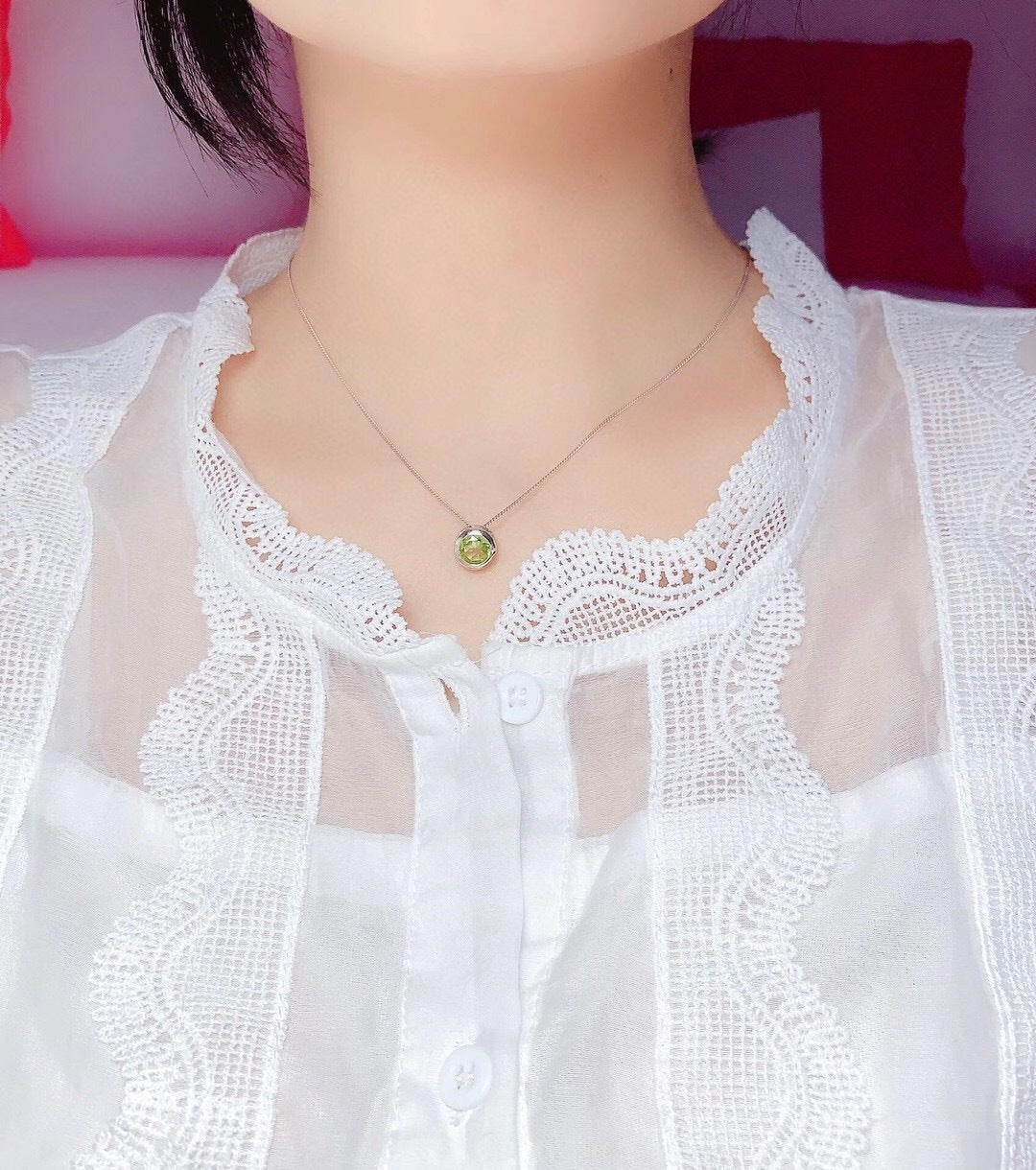 Close-up of a person wearing a white lace top with a necklace featuring a green pendant.
