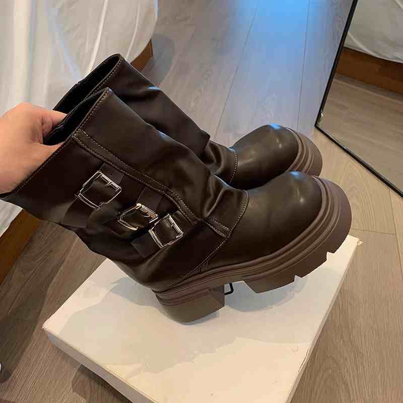 Brown leather boots with buckles held by a hand on a white surface.
